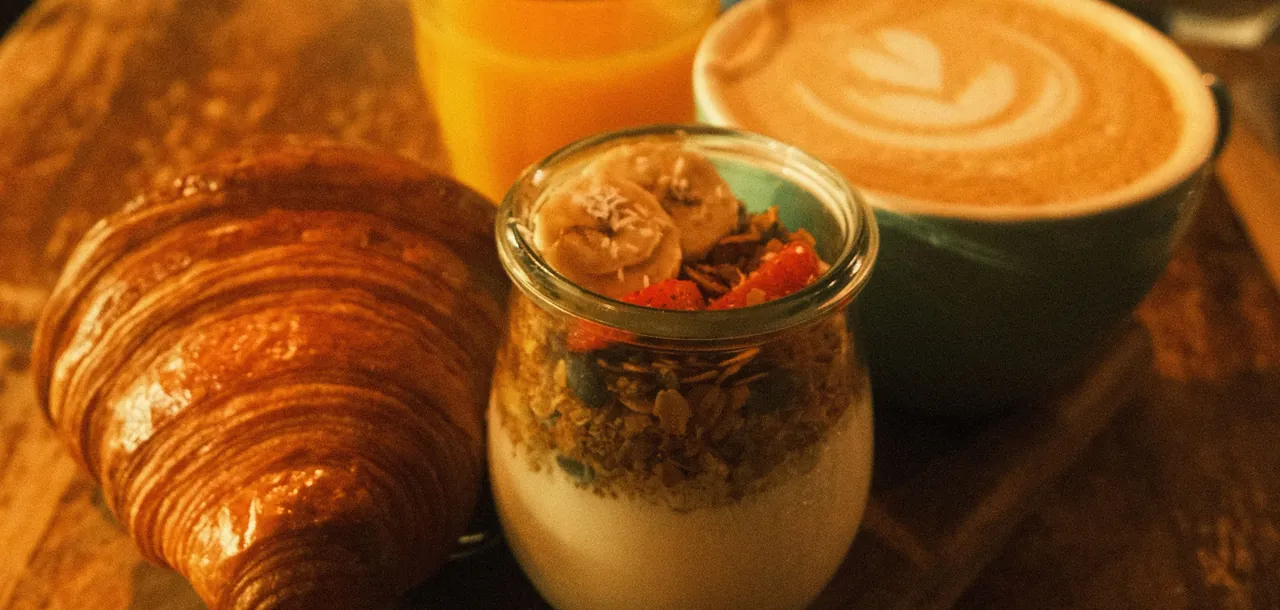 Desayuno servido con croissant, jugo de naranja, yogur con granola y fruta, y un café con arte en la espuma